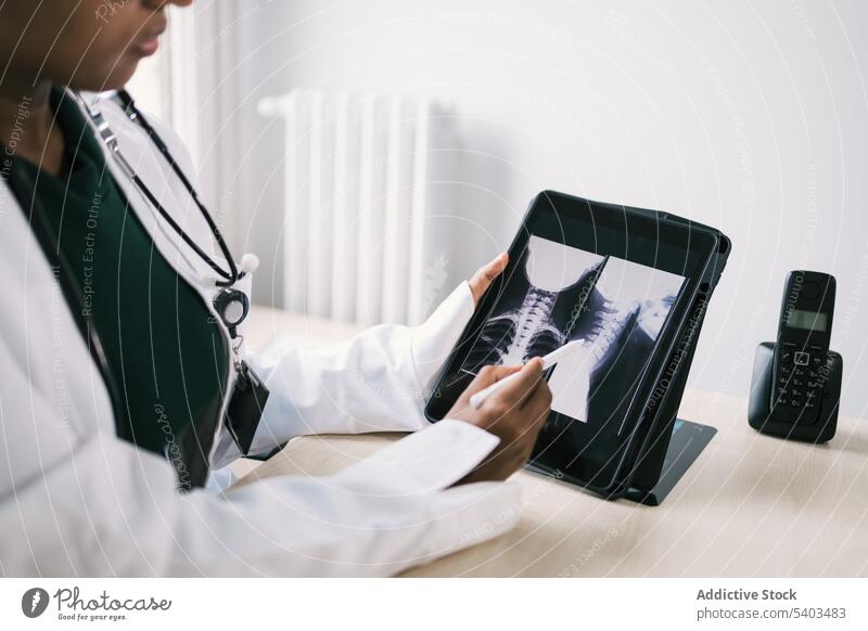 Crop schwarzer Arzt sitzt am Tisch und überprüft Röntgenstrahlen auf Tablette Frau Uniform Stethoskop Röntgenbild Spezialist professionell Raum ethnisch