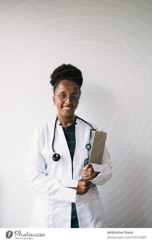 Glücklicher ethnischer Arzt, der mit einem Klemmbrett an der Wand steht Frau Uniform positiv Lächeln Zwischenablage professionell Stethoskop Spezialist Beruf