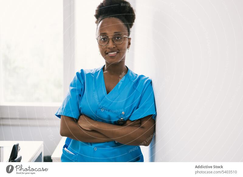 Lächelnde schwarze Frau mit Brille steht an der Wand Arzt Uniform positiv die Hände gekreuzt professionell Glück froh anlehnen jung Afroamerikaner ethnisch