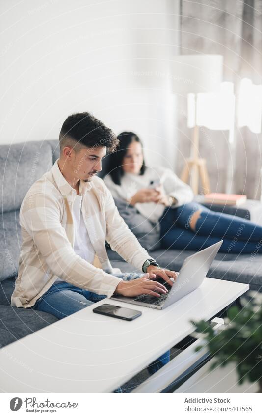Junger Mann arbeitet auf dem Sofa mit Laptop in der Nähe einer Frau mit Mobiltelefon im Wohnzimmer Appartement brünett Kaukasier Computer Paar Entertainment
