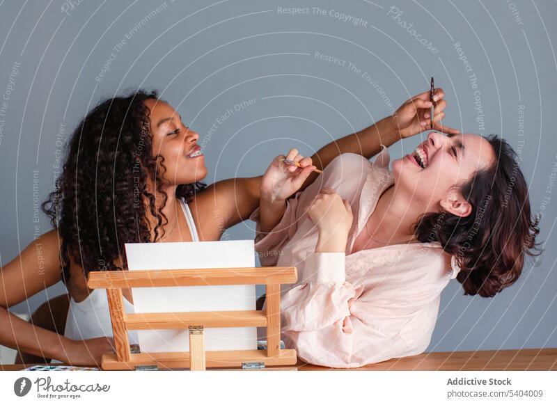 Seitenansicht von fröhlichen multiethnischen Freundinnen, die zu Hause Gesichter bemalen lesbisch Paar Farbe Menschliches Gesicht Pinselblume Zusammensein