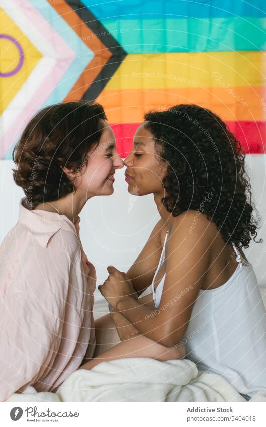 Glückliches lesbisches Paar vor Regenbogenflagge sitzend Frauen Lächeln Fahne lgbt Berührungsnase Augen geschlossen Decke jung Liebe Homosexualität vielfältig