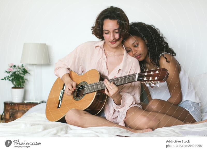 Konzentrierte junge kaukasische Frau spielt Gitarre, während ihre afroamerikanische Freundin den Kopf auf die Schulter stützt und die Musik genießt lesbisch