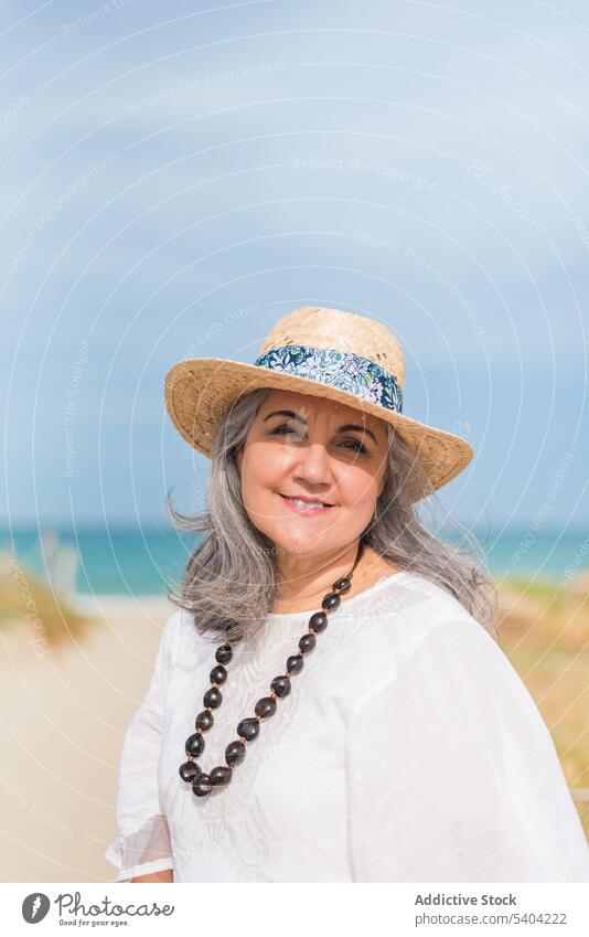 Lächelnde ältere Dame am sonnigen Strand Frau Sommer Meeresufer Küste Porträt Inhalt weißes Kleid reisen Urlaub Seeküste gealtert Stil Ufer Strohhut idyllisch
