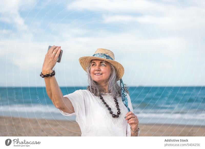 Reife Frau mit Hut nimmt Selfie auf Smartphone in der Nähe von Meer benutzend Strand Sand Küste Strohhut Urlaub Sommer reif Lebensmitte Apparatur Gerät Mobile