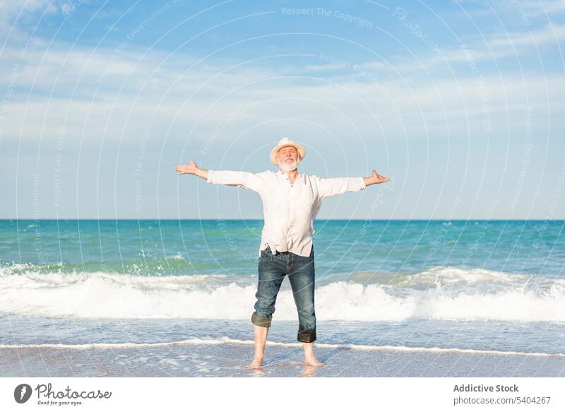 Mann mittleren Alters mit Hut steht am Strand mit ausgestreckten Armen Strohhut Augen geschlossen Barfuß Meerwasser ausdehnen Erholung genießen Urlaub männlich