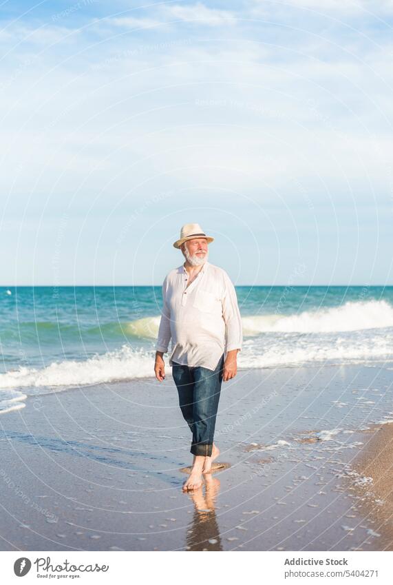 Älterer Mann mit Hut geht am Sandstrand in der Nähe von Meerwasser spazieren Spaziergang Strand Ufer Barfuß Strohhut wolkig Blauer Himmel Urlaub männlich reif