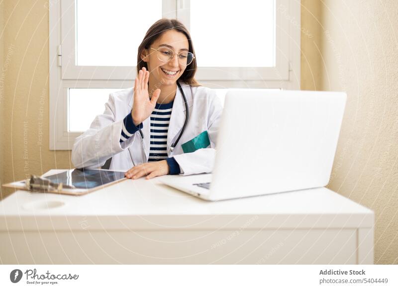 Gut gelaunter Arzt bei einer Online-Videosprechstunde Frau online Videoanruf Berater Laptop Wellenhand professionell Video-Chat Gruß gestikulieren konsultieren