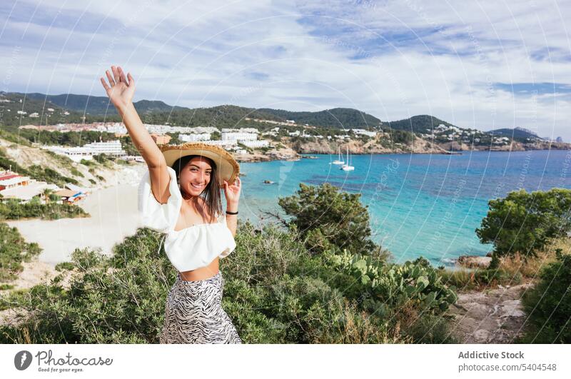 Glückliche Frau an einem tropischen Urlaubsort schaut mit erhobenem Arm in die Kamera Resort Sommer MEER Hut Lächeln Balearen Tourismus reisen Meereslandschaft
