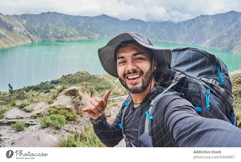 Fröhlicher Reisender mit Rucksack und Hut auf einem Hügel stehend Mann Wanderer Selfie Berge u. Gebirge Lächeln Trekking heiter Ausflug männlich jung Abenteuer