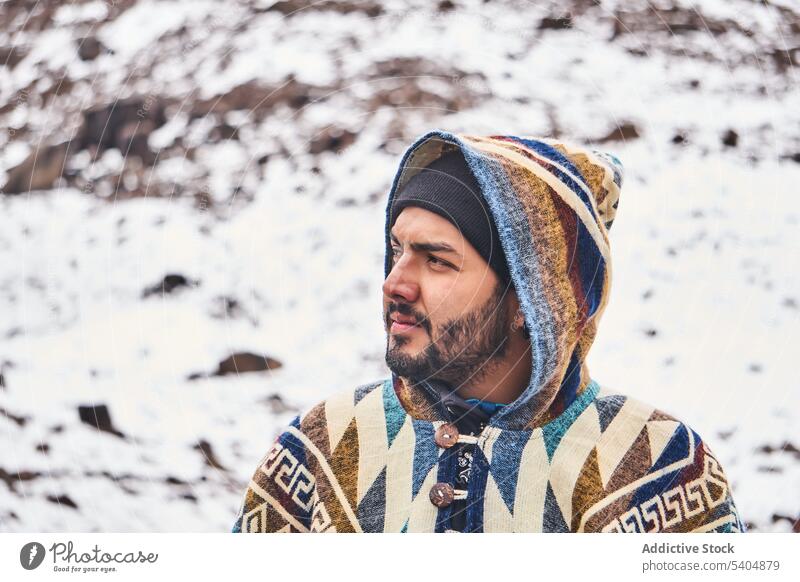 Nachdenklicher junger Mann mit Mütze und Kapuze auf verschneitem Terrain stehend Porträt Berge u. Gebirge kalt Winter Schnee Poncho männlich Vollbart Natur