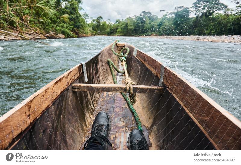 Anonymer Mann in schwarzen Schuhen sitzt auf einem traditionellen Boot, das auf einem Fluss treibt Wasser Baum Schwimmer hölzern Mitfahrgelegenheit Landschaft