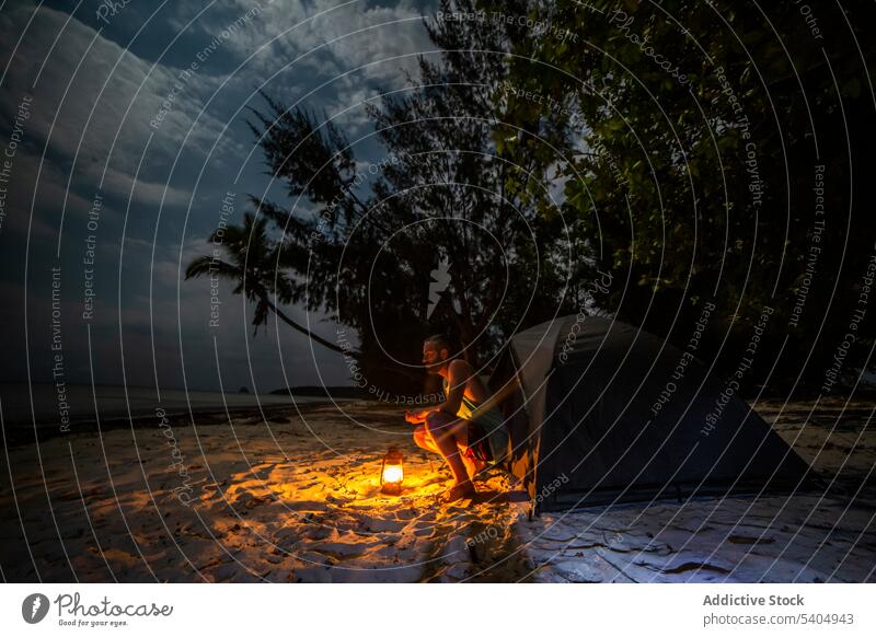 Mann sitzt in der Nähe von Zelt am Sandstrand bei Sonnenuntergang Nacht Lampe Abend Dämmerung MEER Natur Baum Küste Ufer friedlich Windstille idyllisch Strand