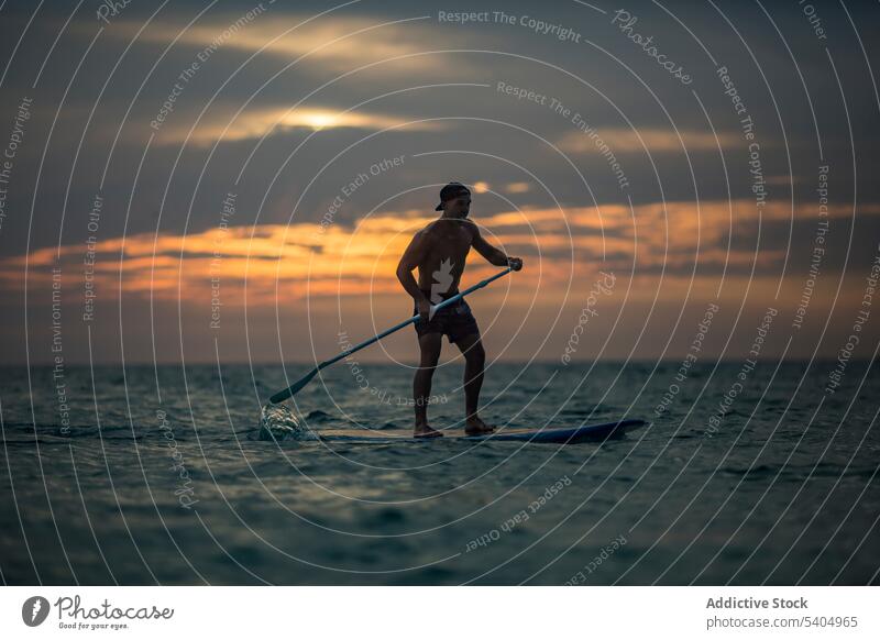 Aktiver männlicher Surfer balanciert auf einem Standup-Paddleboard im Meer Mann aufstehen Paddelbrett MEER Sport passen Sonnenuntergang Natur Aktivität wolkig