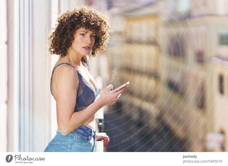 Junge Frau in BH und Jeans mit lockigem Haar beim Surfen auf dem Smartphone krause Haare Unterwäsche Frisur positiv Dessous Balkon Sommer ruhen Sonne heimwärts