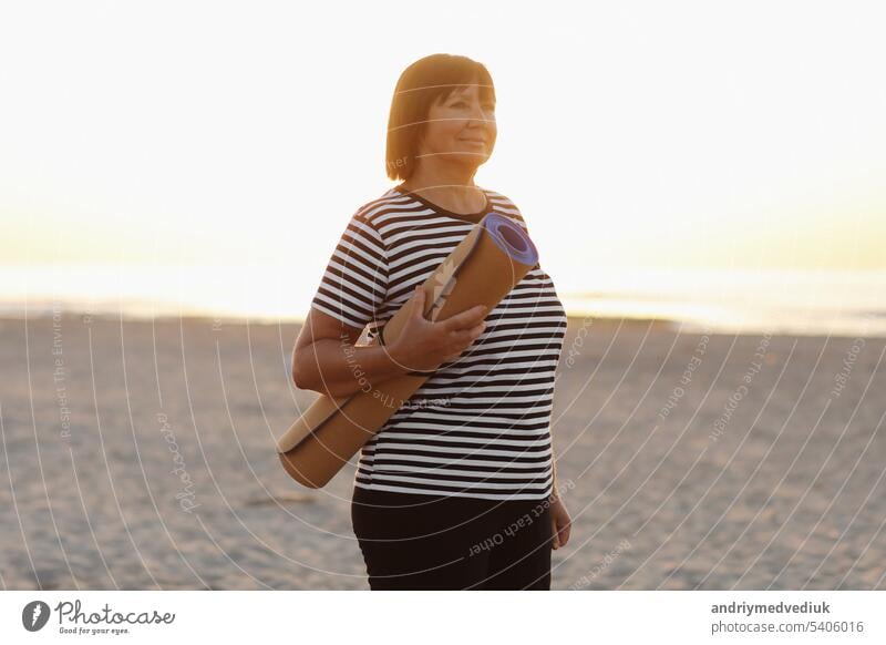 Reife Frau hält eine Sportmatte und bereitet sich darauf vor, Yoga im Freien am Strand zu praktizieren. Glückliche reife übergewichtige Frau, die am Meer trainiert. kopieren Raum. Meditation, Yoga und Entspannung Konzept.