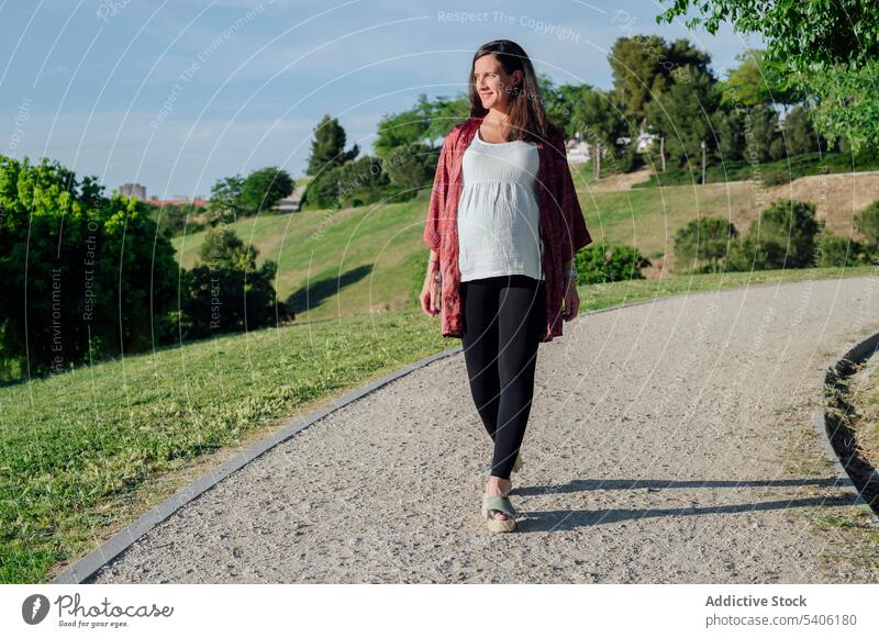 Glückliche schwangere Frau geht im Park spazieren erwarten Bauch Babybauch mütterlich Spaziergang schlendern grün Lächeln Schwangerschaft Kinderkriegen pränatal