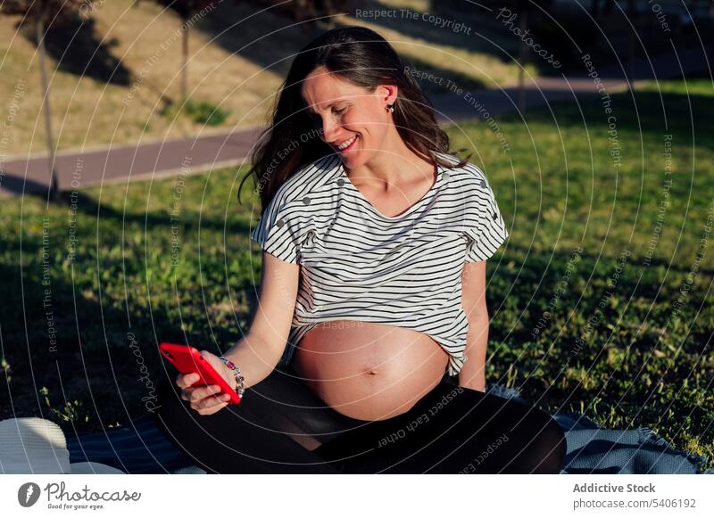 Schwangere Frau mit Smartphone im Park schwanger erwarten Bauch Lotus-Pose Mobile Decke Unterlage Kinderkriegen mütterlich pränatal vorwegnehmen Achtsamkeit