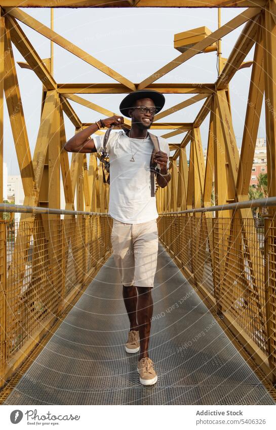 Fröhlicher schwarzer Mann, der lächelnd auf einer Fußgängerbrücke im Park geht Spaziergang Brücke trendy Lächeln Tourist positiv Stil Großstadt Glück männlich