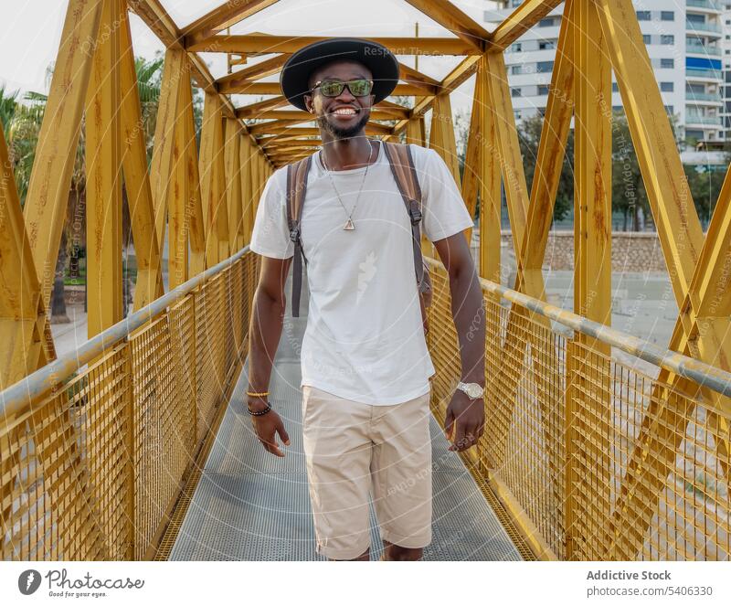 Lächelnder schwarzer Mann, der auf einem Steg im Park geht Spaziergang Brücke trendy Tourist positiv Stil Großstadt Glück männlich jung Afroamerikaner ethnisch