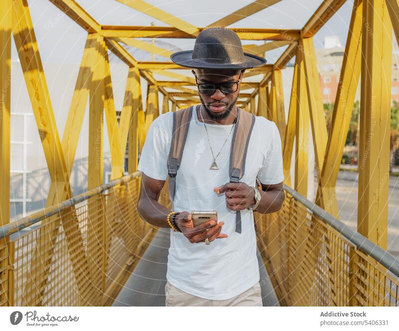 Trendy schwarzer Mann mit Telefon zu Fuß auf Brücke trendy Tourist Smartphone benutzend Spaziergang schlendern Großstadt Straße Rucksack Gerät Apparatur Mobile
