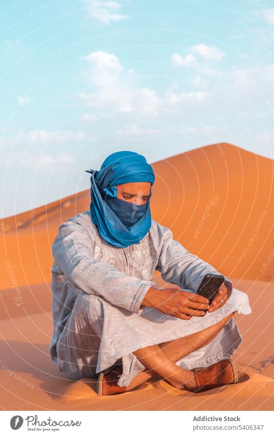 Muslimischer Mann in traditioneller Kleidung, der in der Wüste ein Mobiltelefon benutzt Berber Smartphone benutzend Sand Düne wüst tuareg Tradition Browsen