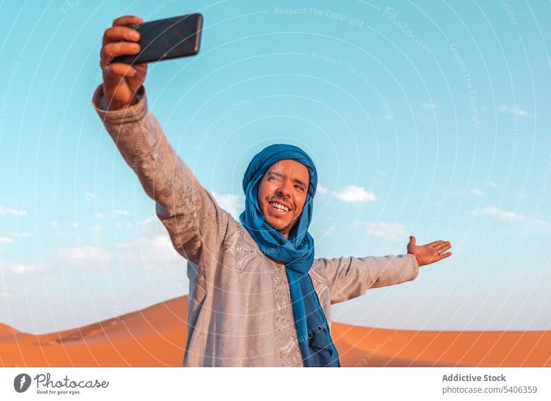 Fröhlicher muslimischer Mann macht Selfie in der Wüste vor blauem Himmel Berber benutzend Smartphone wüst heiter Turban Tradition Lächeln tuareg Marokko reisen