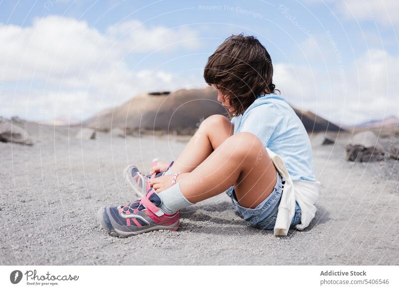 Unbekanntes Kind, das auf trockenem Boden sitzt und sich die Schnürsenkel bindet Krawatte Turnschuh Schuhbänder trocknen Aktivität Sommer Blauer Himmel