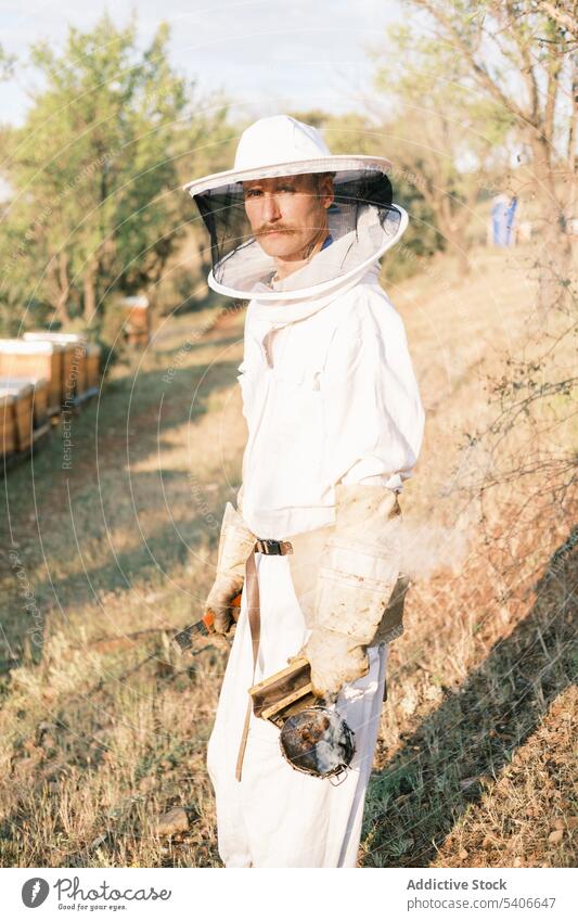 Männlicher Imker mit Raucher bei der Arbeit im Bienenstock Mann Raucherin Bienenkorb Werkzeug Ackerbau behüten professionell prüfen Gerät Uniform Bauernhof