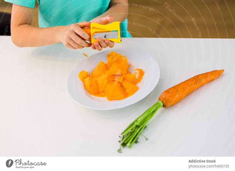 Anonymer Junge am Tisch stehend mit frischer Karotte vor verschwommener Küche Kind Scheibe Möhre Teller Hilfsbereitschaft Anspitzer beschäftigt heimwärts