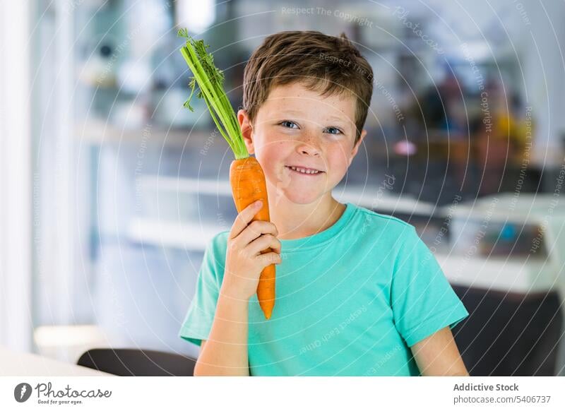 Lächelnder Junge mit Karotte vor verschwommener Küche Möhre reif gesunde Ernährung Gesundheit Kind niedlich bezaubernd positiv lecker geschmackvoll süß