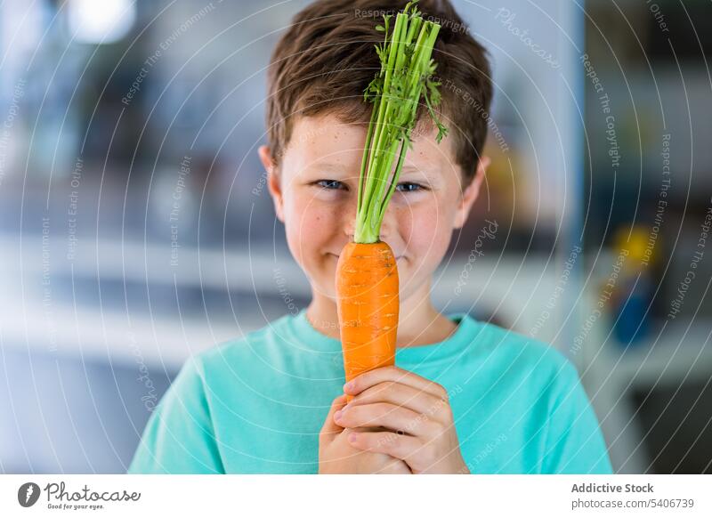 Lächelnder Junge zeigt Karotte gegen unscharfe Küche Möhre reif gesunde Ernährung Gesundheit Kind niedlich bezaubernd positiv lecker geschmackvoll süß organisch