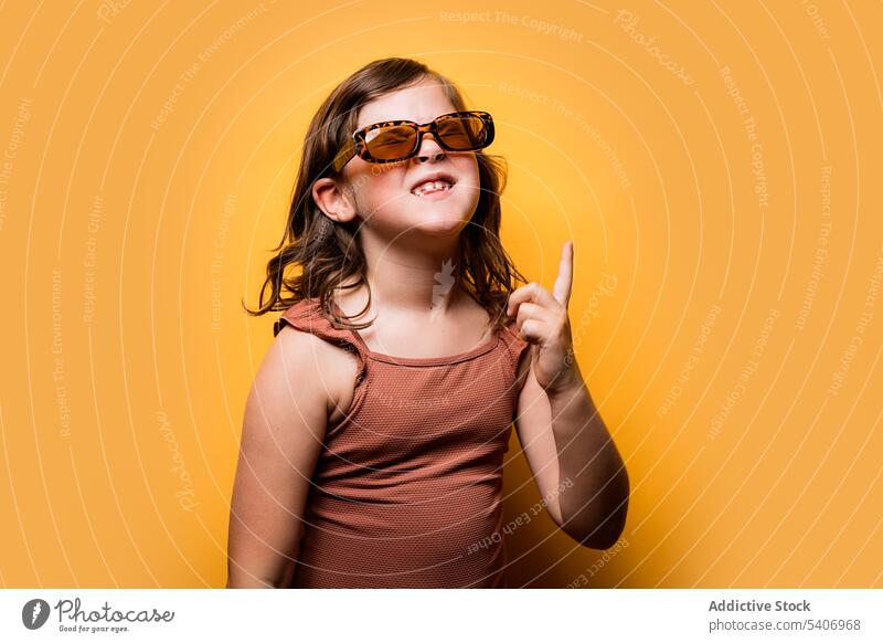 Freudiges kleines Kind in Sommerkleidung, das in einem orangefarbenen Studio nach oben zeigt Mädchen nach oben zeigen Augen geschlossen Glück heiter lustig