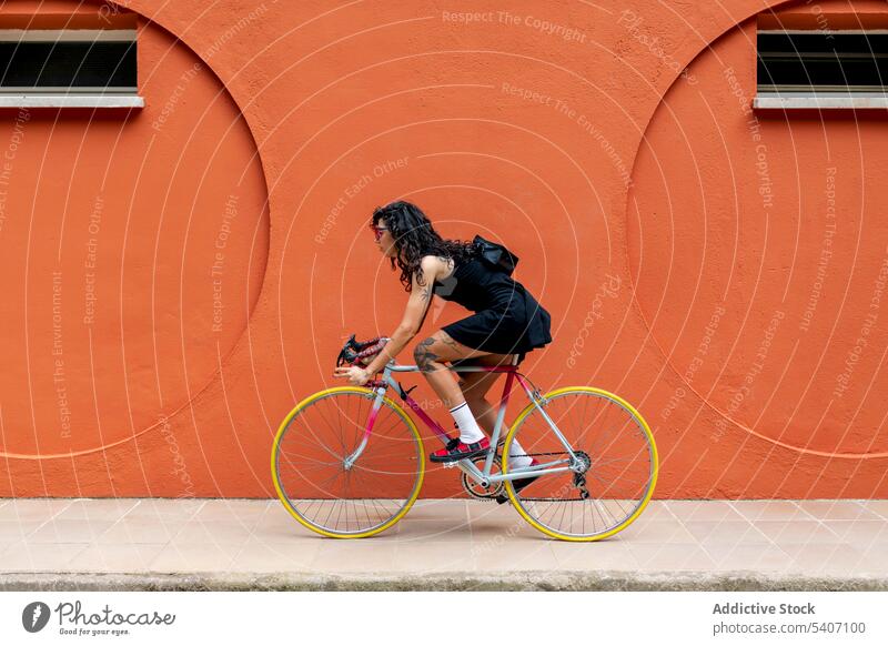 Junge ethnische Frau mit Handtasche auf dem Fahrrad auf dem Bürgersteig in der Nähe der orangefarbenen Mauer Straße Mitfahrgelegenheit Wand Gebäude Turnschuh