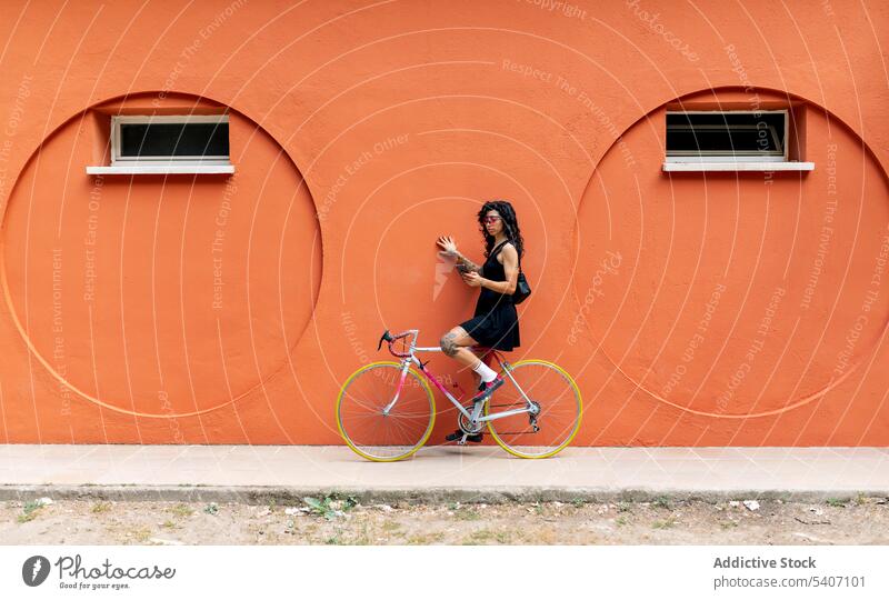 Junge ethnische Frau, die auf einem Fahrrad sitzt und in der Nähe einer orangefarbenen Wand auf ihrem Smartphone surft Straße benutzend Bürgersteig online
