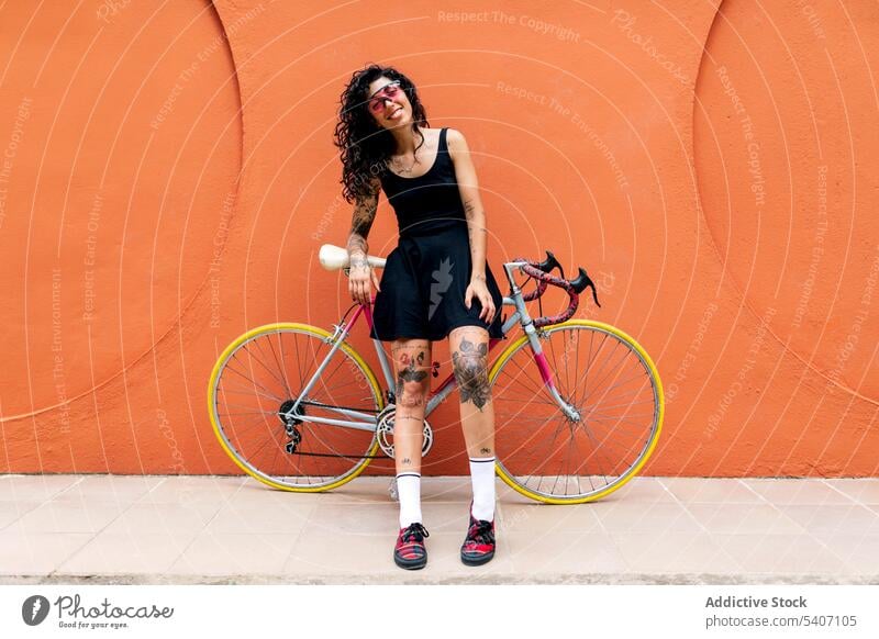 Junge ethnische Frau lehnt an einem Fahrrad Bürgersteig Straße orange Wand Gebäude Verkehr Sommer jung hispanisch Lächeln Straßenbelag schlendern Sonnenbrille
