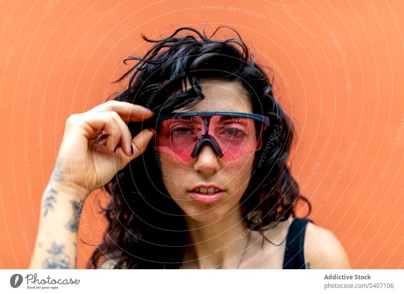Stilvolle junge ethnische Frau berührt mit der Hand eine Sonnenbrille im Tageslicht Porträt Lächeln Glück orange Wand Tattoo Sommer hispanisch tagsüber feminin