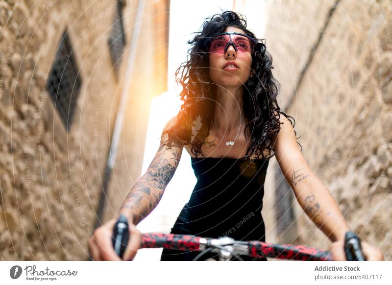Stilvolle tätowierte junge ethnische Frau steht in einer engen Straße mit Fahrrad selbstbewusst Backsteinwand Gebäude Vorschein Verkehr Sommer hispanisch
