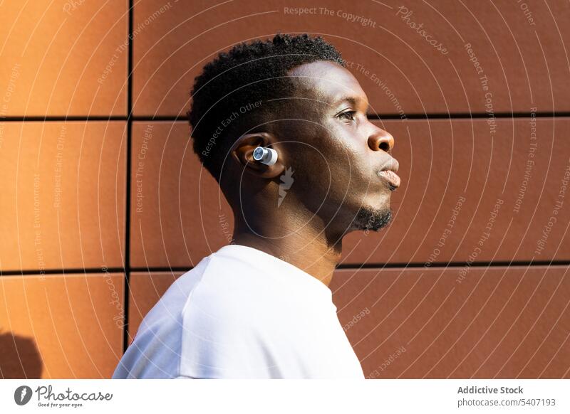 Ernster junger schwarzer Mann an der Wand stehend zuhören Porträt Musik ernst Kopfhörer T-Shirt besinnlich männlich echte drahtlose tws Afroamerikaner