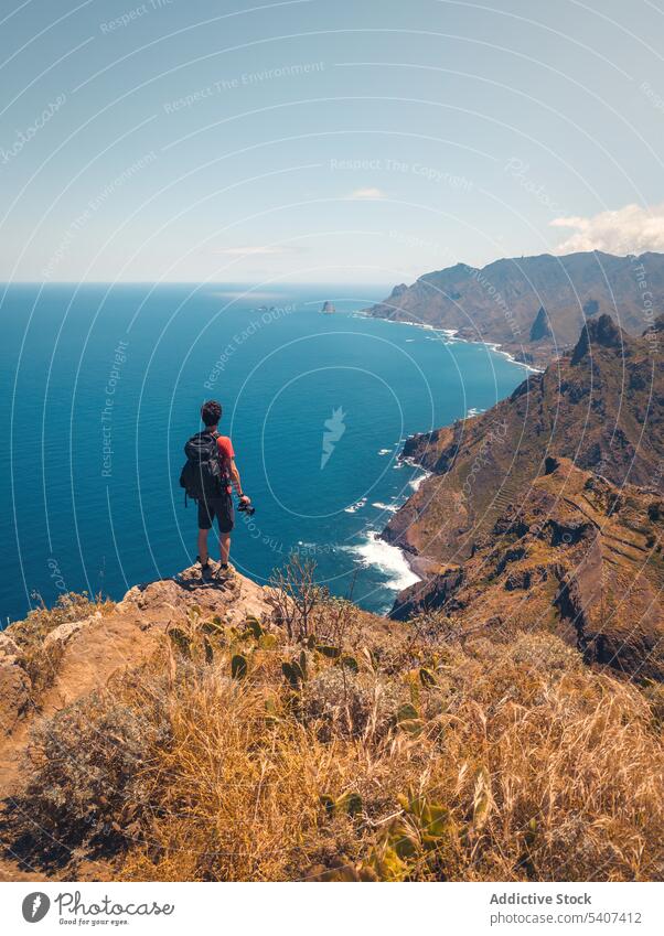 Junger Mann mit Rucksack auf Klippe stehend mit Kamera bewundernd Meereslandschaft Berge u. Gebirge Reisender Wanderer Natur MEER Landschaft Abenteuer männlich