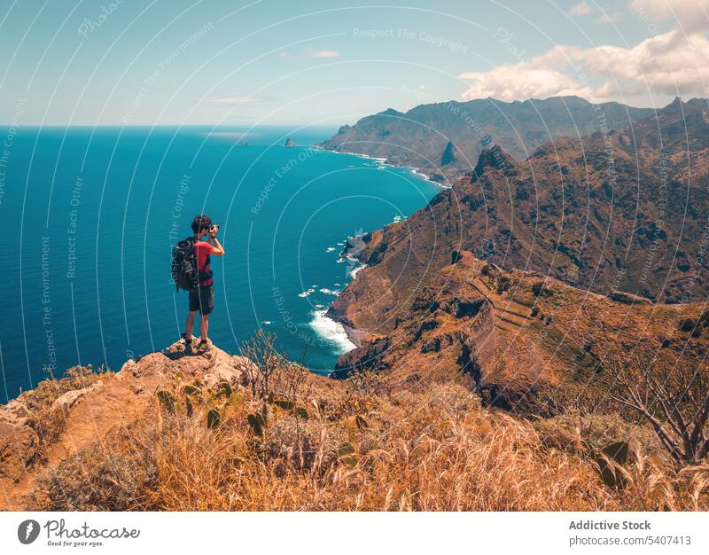 Junger Mann mit Rucksack auf einer Klippe stehend und mit Kamera fotografierend Berge u. Gebirge Reisender Wanderer Natur MEER Landschaft Abenteuer männlich