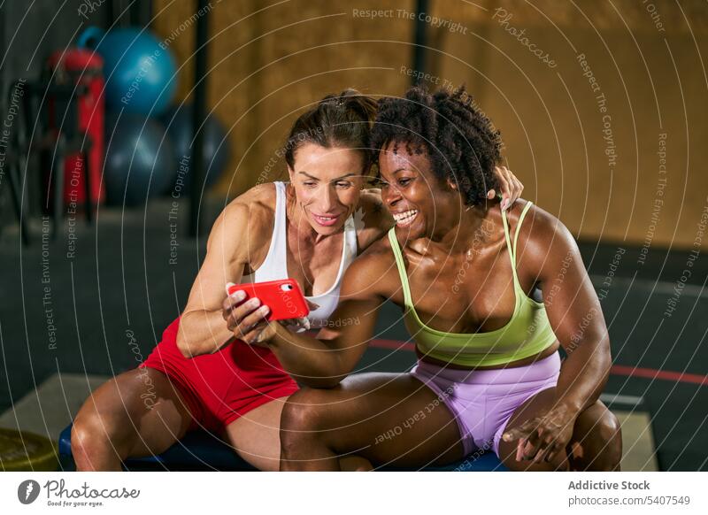 Fröhliche, vielseitige Sportlerinnen sehen sich ein Video auf ihrem Smartphone an benutzend zuschauen lustig teilen Pause Training Fitnessstudio Lächeln heiter