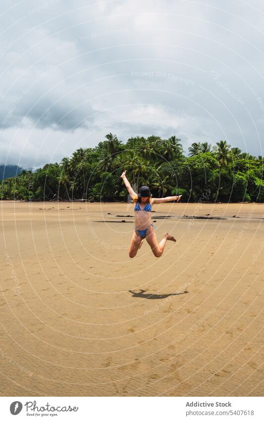 Unbekümmerte Frau springt am Sandstrand Resort Sonnenlicht springen Freiheit Arme hochgezogen sorgenfrei Strand Ufer Spaß haben Freude aufgeregt Energie jung