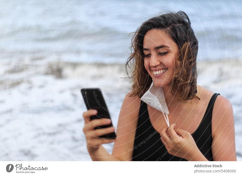 Fröhliche Reisende, die mit ihrem Smartphone in der Nähe des wogenden Meeres plaudert Reisender plaudernd Internet online heiter Mundschutz COVID benutzend