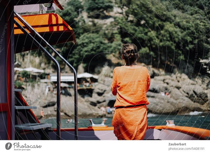 Touristin im orangen Kleid steht auf Boot in einer Bucht an der felsigen Küste an der italienischen Riviera Italien Reling beobachten reisen sonnig Wasser Meer