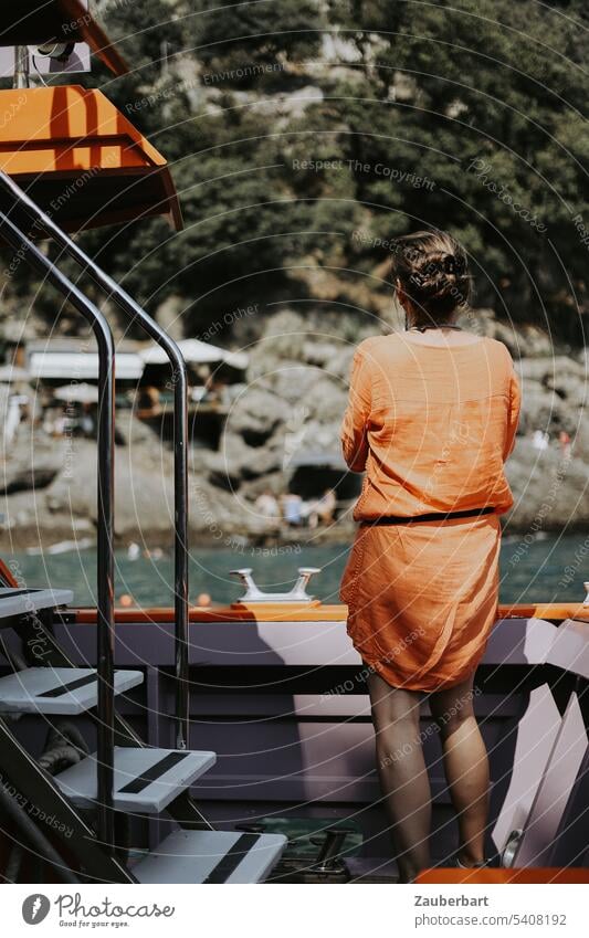 Touristin im orangen Kleid steht auf Boot in einer Bucht an der felsigen Küste an der italienischen Riviera Italien Reling beobachten reisen sonnig Wasser Meer