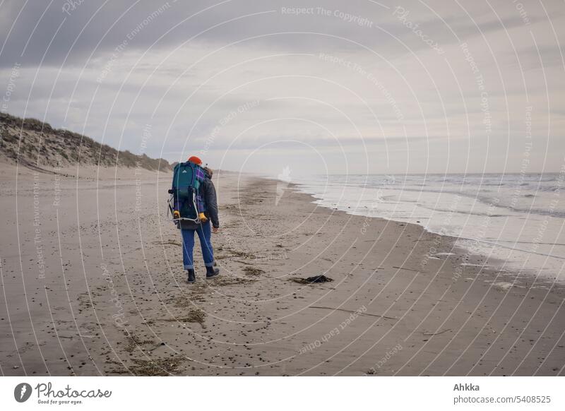 Frau mit Kind im Rucksack spaziert an einem herbstlichen Strand Kindheitserinnerung spazieren Stranddüne Wasser Meer Wellen Himmel weite Ausflug