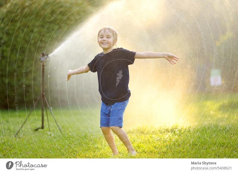 Lustiger kleiner Junge spielt mit Garten-Sprinkler im sonnigen Stadtpark. Grundschulkind lacht, springt und hat Spaß mit Sprühwasser. Sommer im Freien Aktivität für Kinder.