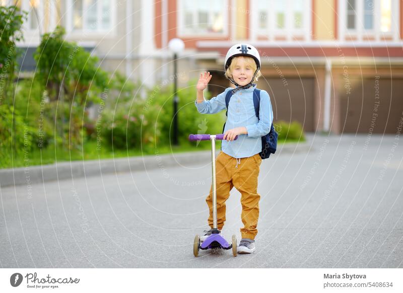 Kleines Kind mit Schutzhelm fährt mit dem Roller zur Schule. Vorschüler Junge winkt Hand sagen hallo. Sicherheit Kinder auf dem Weg zur Schule. Zurück zu Schule Konzept.