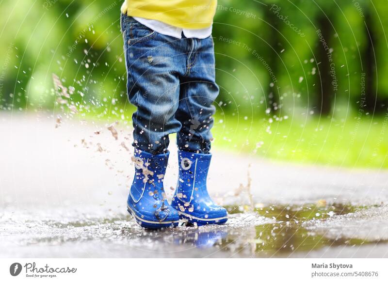 Ein Kleinkind, das an einem Sommer- oder Herbsttag in ein Wasserbecken springt. Baby Spaß springen Pfütze im Freien Kind Junge Regen Stiefel Spielzeit platschen